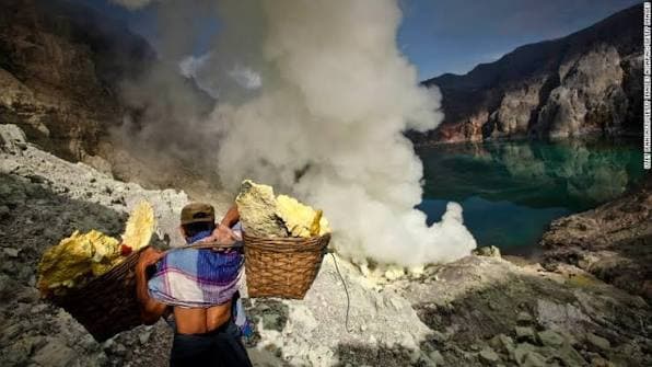 A sulphur miner carrying two loaded baskets on a shoulder pole out of the Ijen crater, sulphurous smoke and the turquoise lake behind