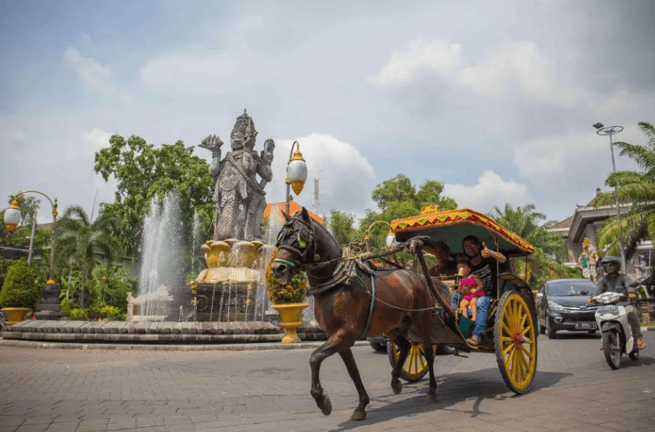 A dokar horse cart carrying passengers past a fountain and stone guardian statue in Surakarta