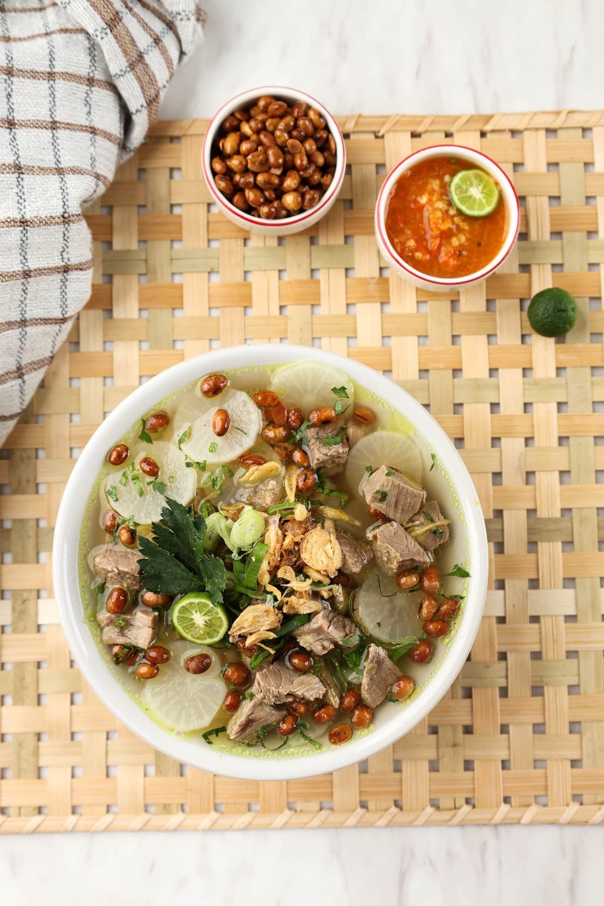 A bowl of soto Bandung, clear beef soup with radish and fried soybeans, seen from above on a woven mat